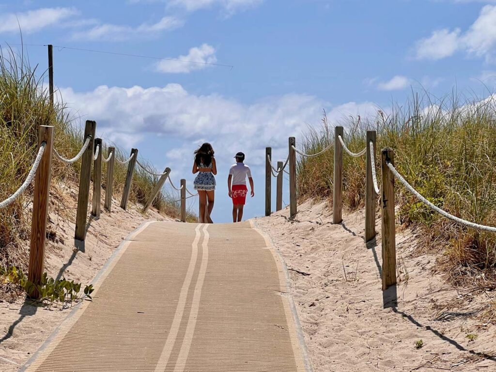 teen girl and boy walking along a sandy path toward the beach, surrounded by dunes and clear skies at Loews Miami Beach."