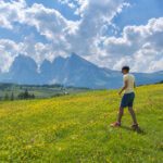 A boy in a yellow shirt walks through a wildflower meadow with the Dolomite mountains and cloudy blue sky in the background.