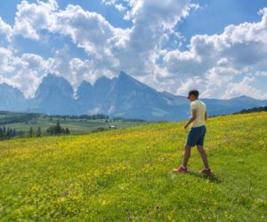 A boy in a yellow shirt walks through a wildflower meadow with the Dolomite mountains and cloudy blue sky in the background.